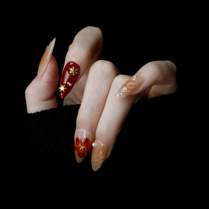 A pair of hands adorned with Elegant Floral Red and Beige Press-On Nails by We Do Nails, featuring intricate red flowers and pearl-gold accents, holds a large black mug. The elongated, almond-shaped nails boast a glossy finish with 3D floral designs against the black background.