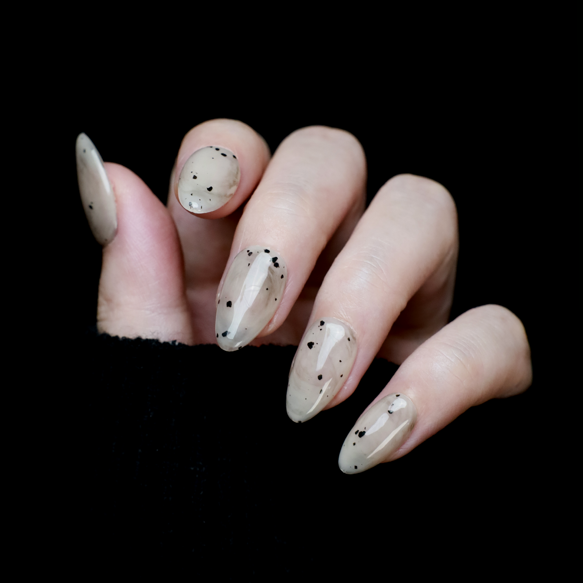 Hands showcasing We Do Nails Marble and Speckled Press On Nails, almond-shaped with a high-gloss shine and scattered speckles, perfectly highlighted against a solid black background.