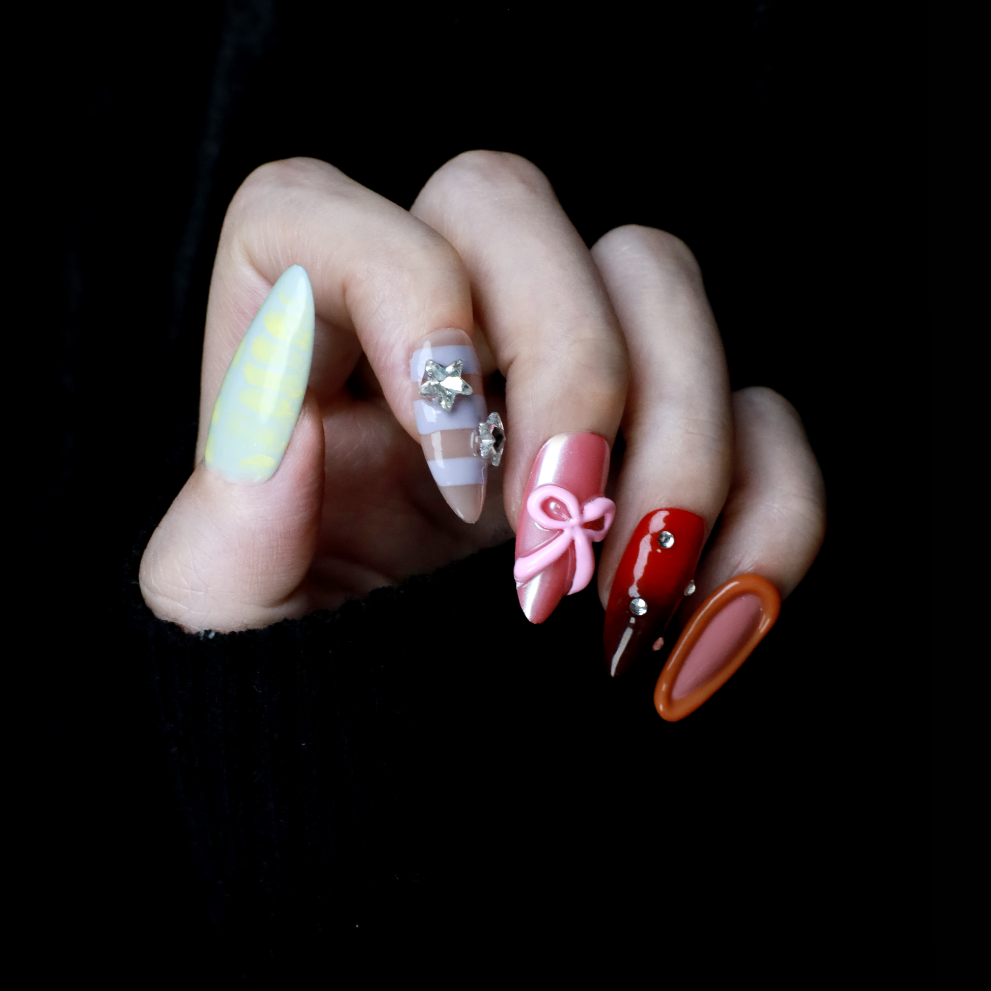 A hand with We Do Nails Candyland Dream Press-On Nails displays playful designs. Features include pastel yellow plaid, clear nails with bows and rhinestones, pink nails with a bow, shiny red with rhinestones, and glossy rust-orange—all set against a dark backdrop.