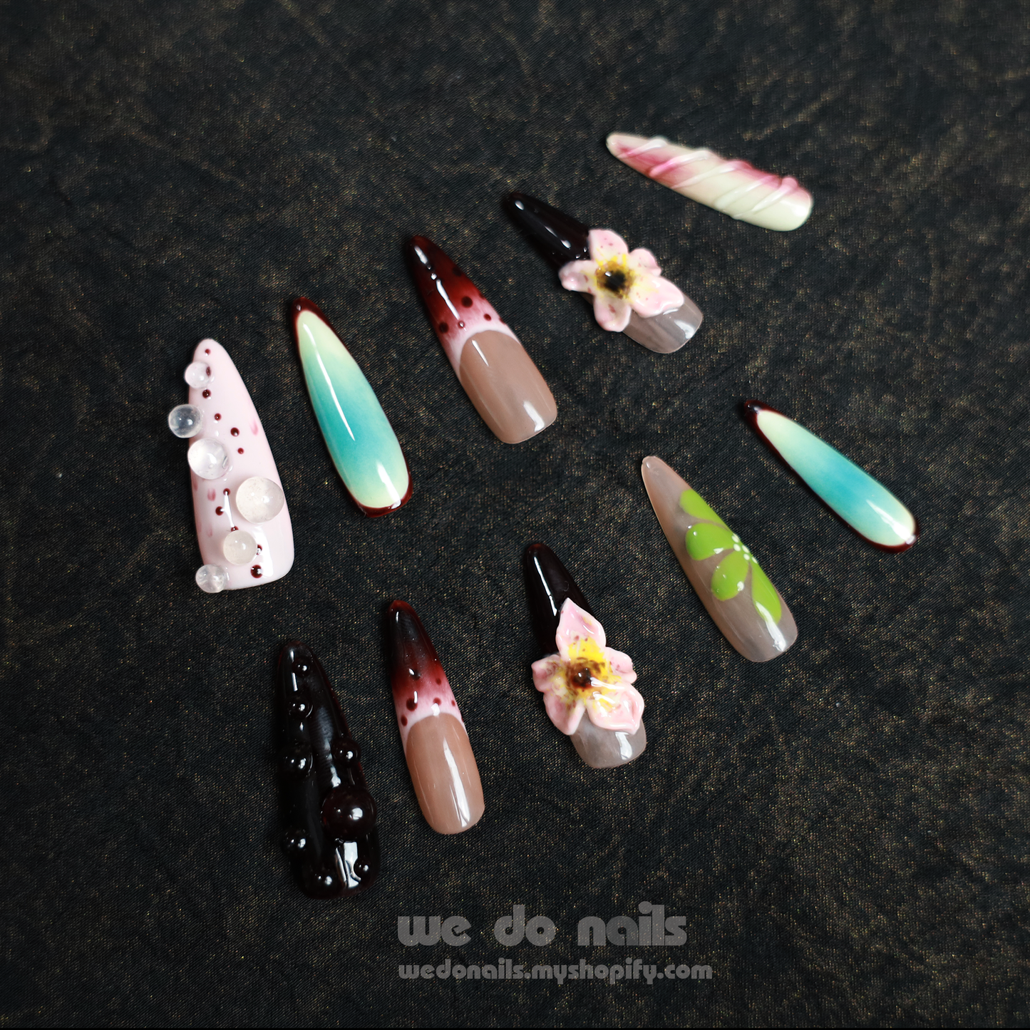 A close-up shows two hands with Artistic Floral Spring Press-On Nails from We Do Nails. The left features floral and gem patterns on black, while the right displays blue ombre with floral tips, all set against a dark backdrop.