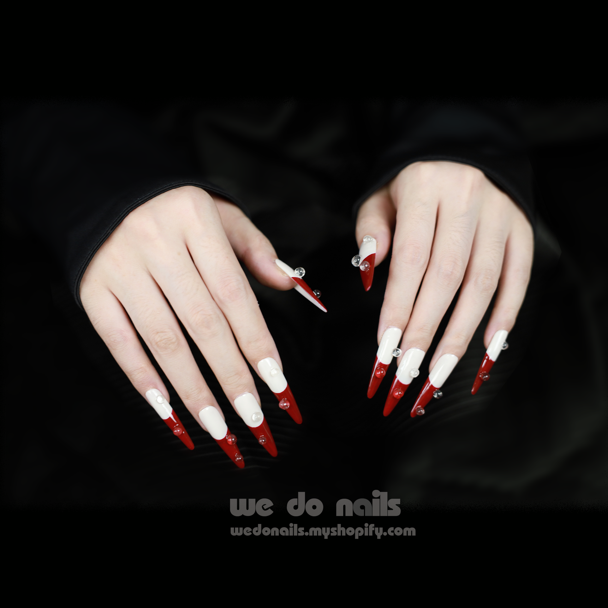 Close-up of two hands with long, pointed Pacific Flame Press-On Nails by We Do Nails, painted half white and half dark red with heart decorations against a black background.
