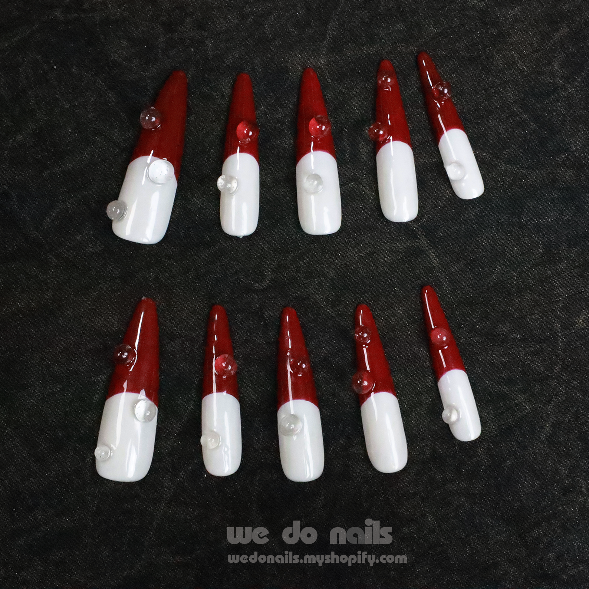 Close-up of two hands with long, pointed Pacific Flame Press-On Nails by We Do Nails, painted half white and half dark red with heart decorations against a black background.