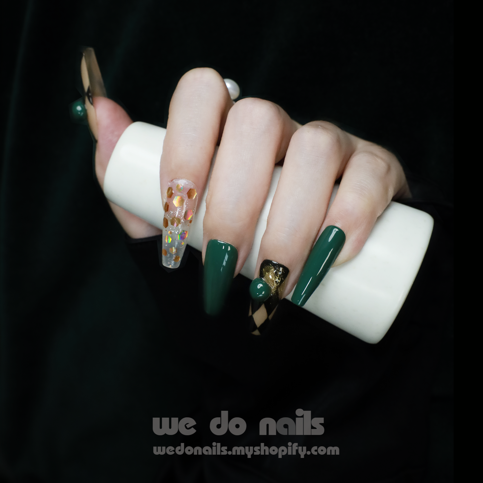 A hand with stylish Green Geometric Press-On Nails by We Do Nails holds a white cylinder. The nails are glossy dark green, accented by a transparent nail with gold flakes. A dark background highlights the detailed nail art reminiscent of an elegant, old-fashioned style.