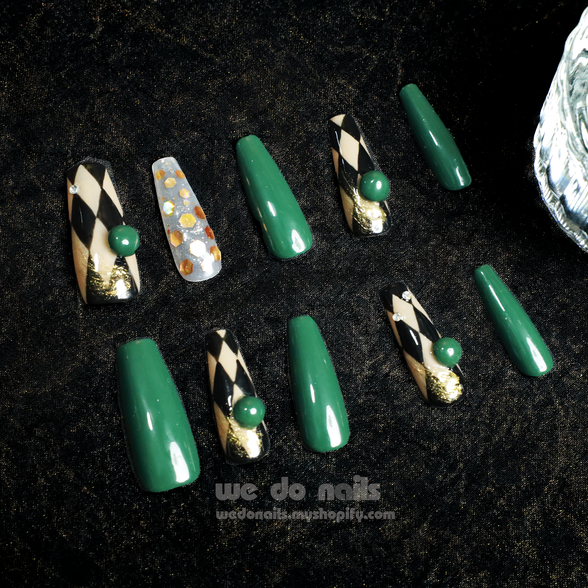 A hand with stylish Green Geometric Press-On Nails by We Do Nails holds a white cylinder. The nails are glossy dark green, accented by a transparent nail with gold flakes. A dark background highlights the detailed nail art reminiscent of an elegant, old-fashioned style.