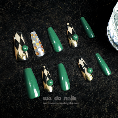 A hand with stylish Green Geometric Press-On Nails by We Do Nails holds a white cylinder. The nails are glossy dark green, accented by a transparent nail with gold flakes. A dark background highlights the detailed nail art reminiscent of an elegant, old-fashioned style.