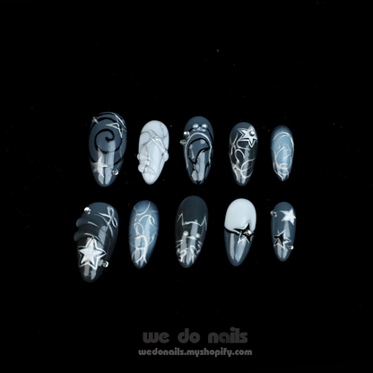 A person holds a white mug with both hands, showcasing long, pointed nails from We Do Nails Midnight Custom Press-On Nails. Star and galaxy designs exude cosmic elegance against a dark background, enhancing the vibrant celestial wonder of the nail art.