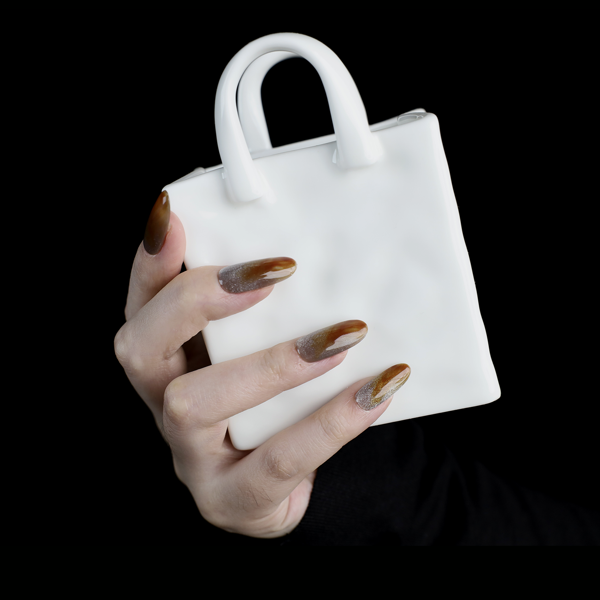 A hand with We Do Nails Elegant Brown Marble Press-On Nails, featuring a subtle metallic chrome accent, holds a simple white mug against a black backdrop.
