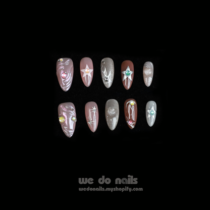 Two hands with long, decorated nails hold a white ceramic mug. Featuring Shiny Star Press-On Nails by We Do Nails, the celestial-inspired manicure showcases flowers, stars, and glitter. Against a dark background, We Do Nails and a URL add allure to this luminous display.