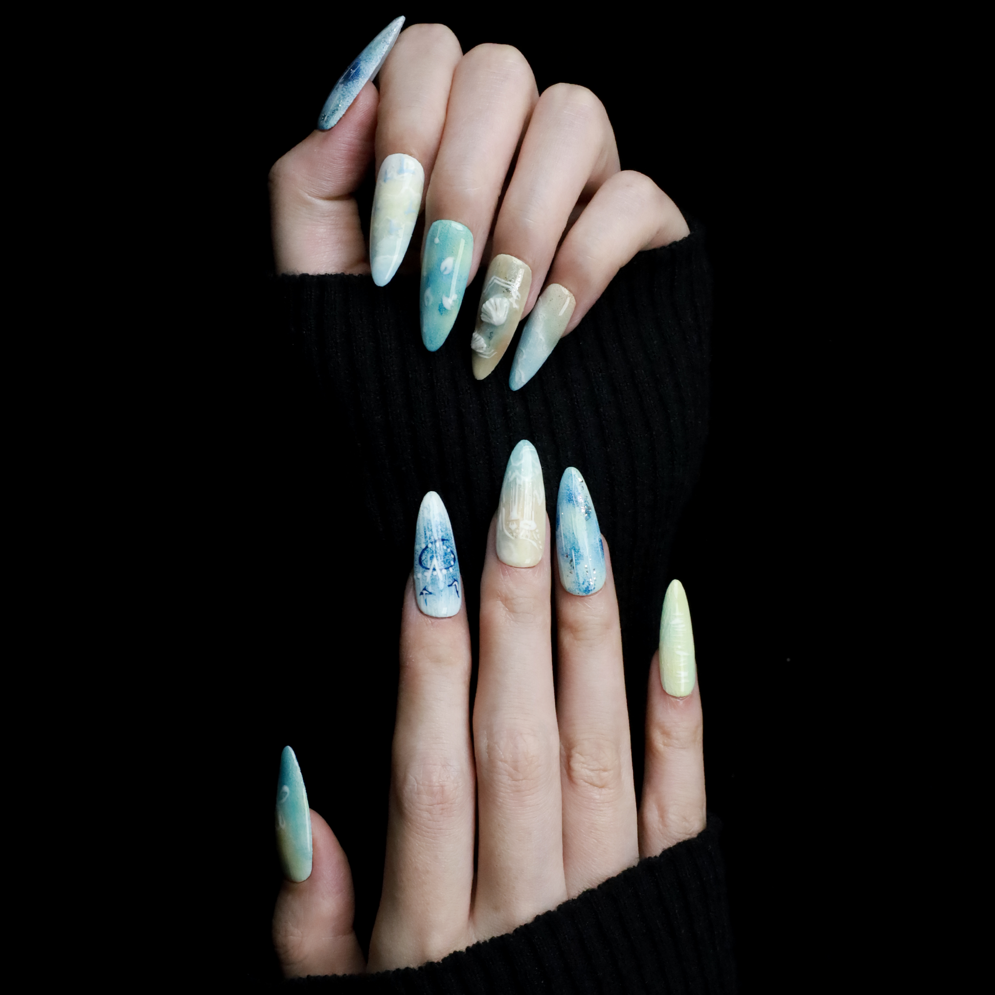 A hand flaunts the elegant Ocean-Inspired Press-On Nails by We Do Nails, featuring long, pointed shapes with ocean-themed designs. The set showcases ocean breeze colors, seashell art, and wave patterns against a dark background to highlight its intricate beauty.