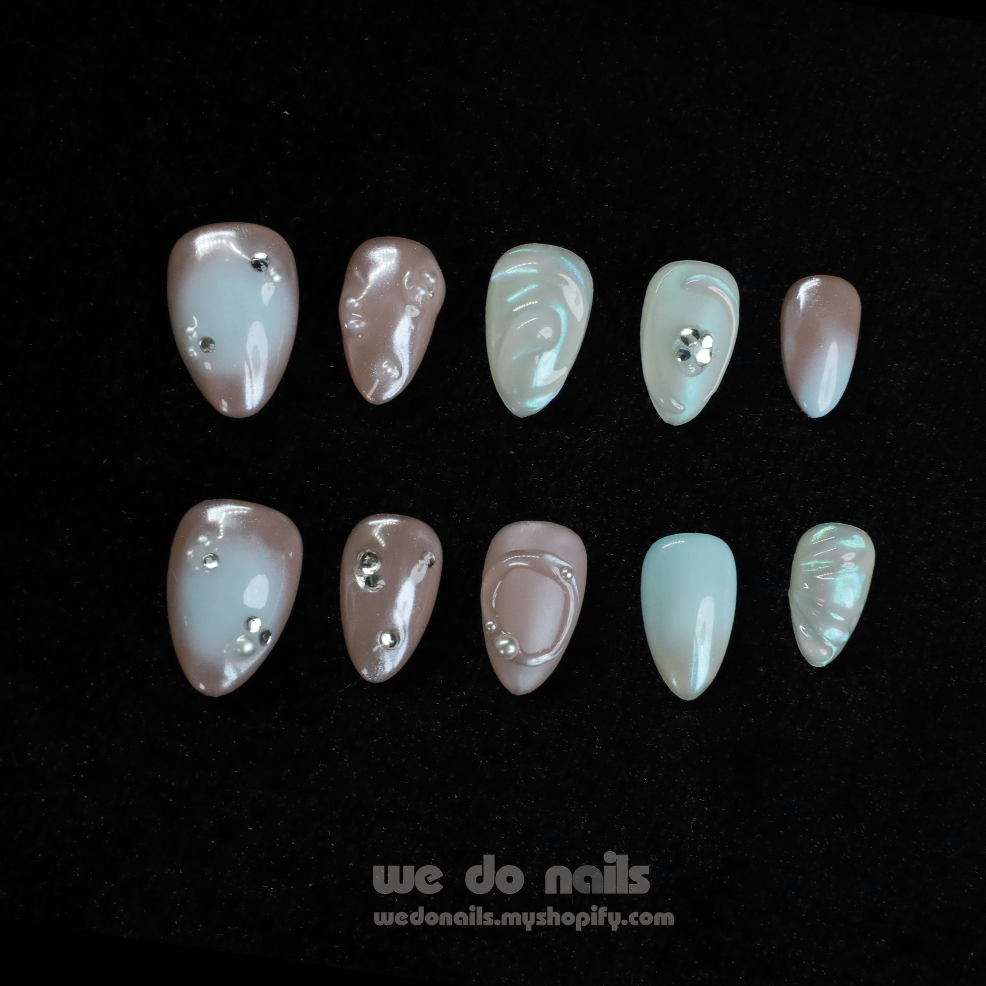 Close-up of hands with Pearl Dreams Press-On Nails by We Do Nails, showcasing manicured nails in small patterns and neutral colors. Subtle pearlescent hues add elegance. Fingers are gently intertwined, wearing a black long-sleeve sweater. We Do Nails text visible on the left.