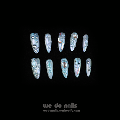Displaying interlocked fingers adorned with We Do Nails Iridescent Bubble Press-On Nails, these long, pointed artificial nails feature a holographic silver and pearl finish, beautifully contrasted by a sleek black garment. Text reads we do nails and wedonails.myshopify.com.