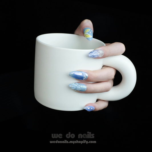 A hand sporting Under The Sea Press-On Nails in blue and silver, resembling mermaid tails, holds a large white mug with a modern design against a dark background. At the bottom, We Do Nails is displayed along with a website link.