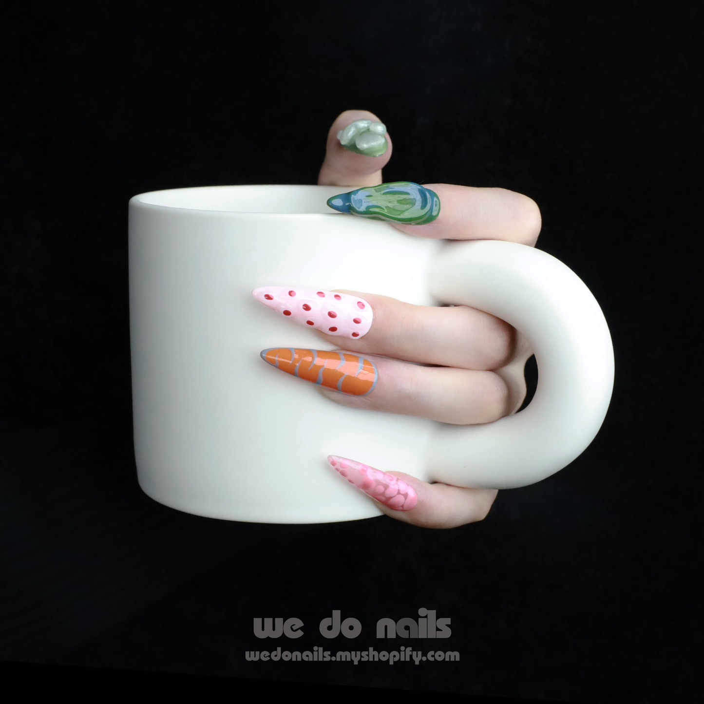 A hand with We Do Nails Tropical Vibe Press-On Nails, featuring a chic mix of pink, orange, and green geometric designs like polka dots and abstract shapes, holds a large white mug. The dark background enhances the trendy look, ideal for beach parties or summer vacations.