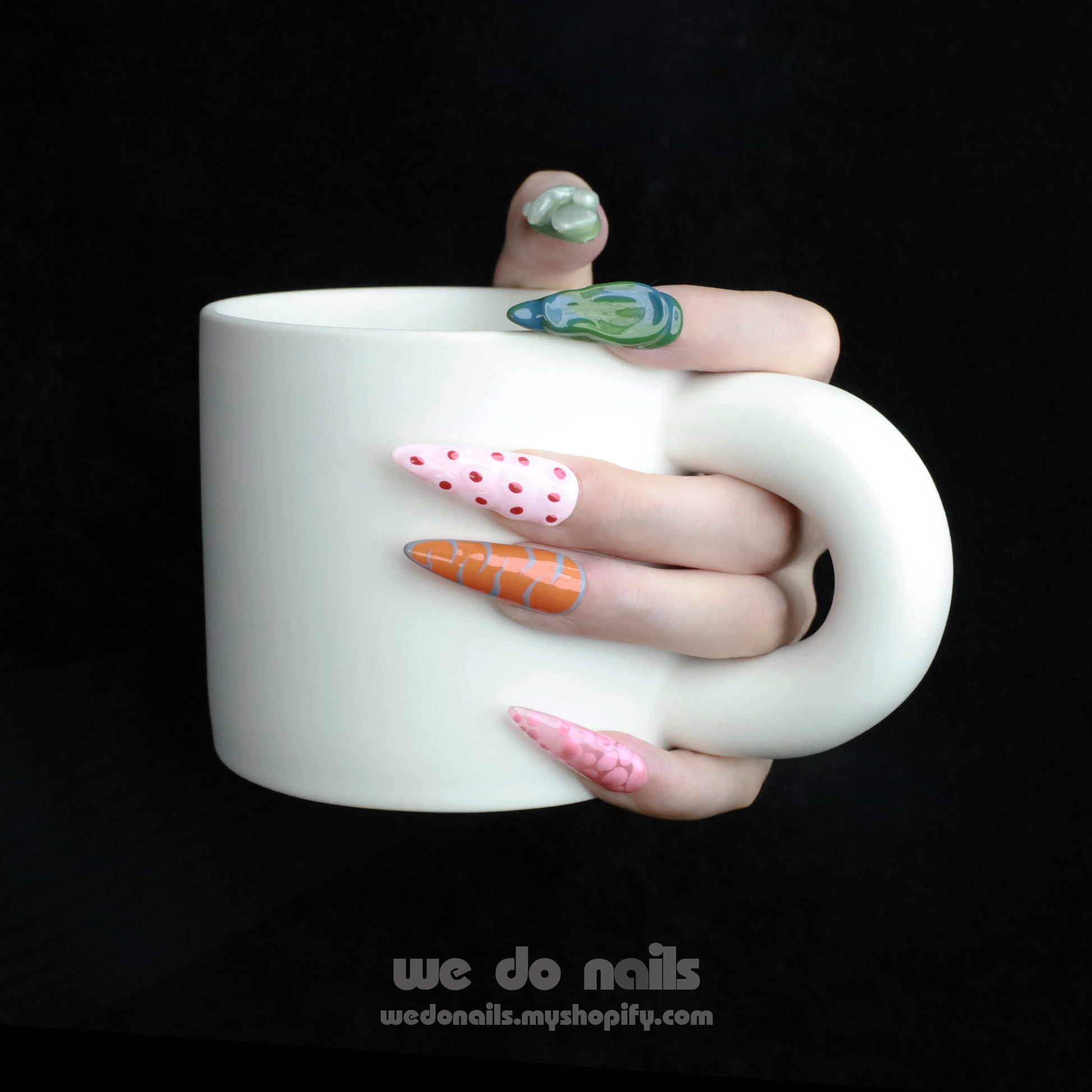 A hand with We Do Nails Tropical Vibe Press-On Nails, featuring a chic mix of pink, orange, and green geometric designs like polka dots and abstract shapes, holds a large white mug. The dark background enhances the trendy look, ideal for beach parties or summer vacations.