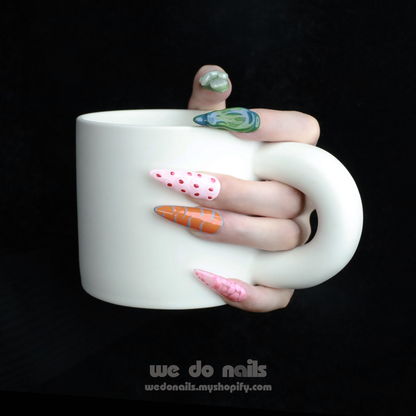 A hand with We Do Nails Tropical Vibe Press-On Nails, featuring a chic mix of pink, orange, and green geometric designs like polka dots and abstract shapes, holds a large white mug. The dark background enhances the trendy look, ideal for beach parties or summer vacations.