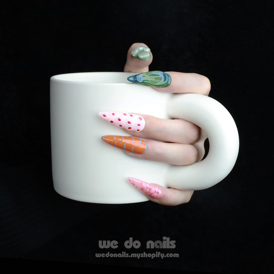 A hand with We Do Nails Tropical Vibe Press-On Nails, featuring a chic mix of pink, orange, and green geometric designs like polka dots and abstract shapes, holds a large white mug. The dark background enhances the trendy look, ideal for beach parties or summer vacations.
