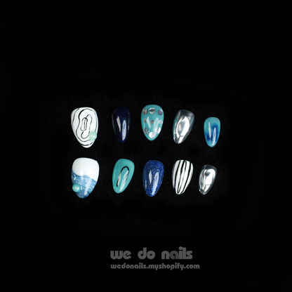 A person in a black sweater displays manicured fingernails with enchanting designs like polka dots, stripes, and abstract patterns. The metallic finishes shine in blue ombre and green hues, featuring Midnight Mystique Press-On Nails by We Do Nails. The text reads we do nails next to a website URL.