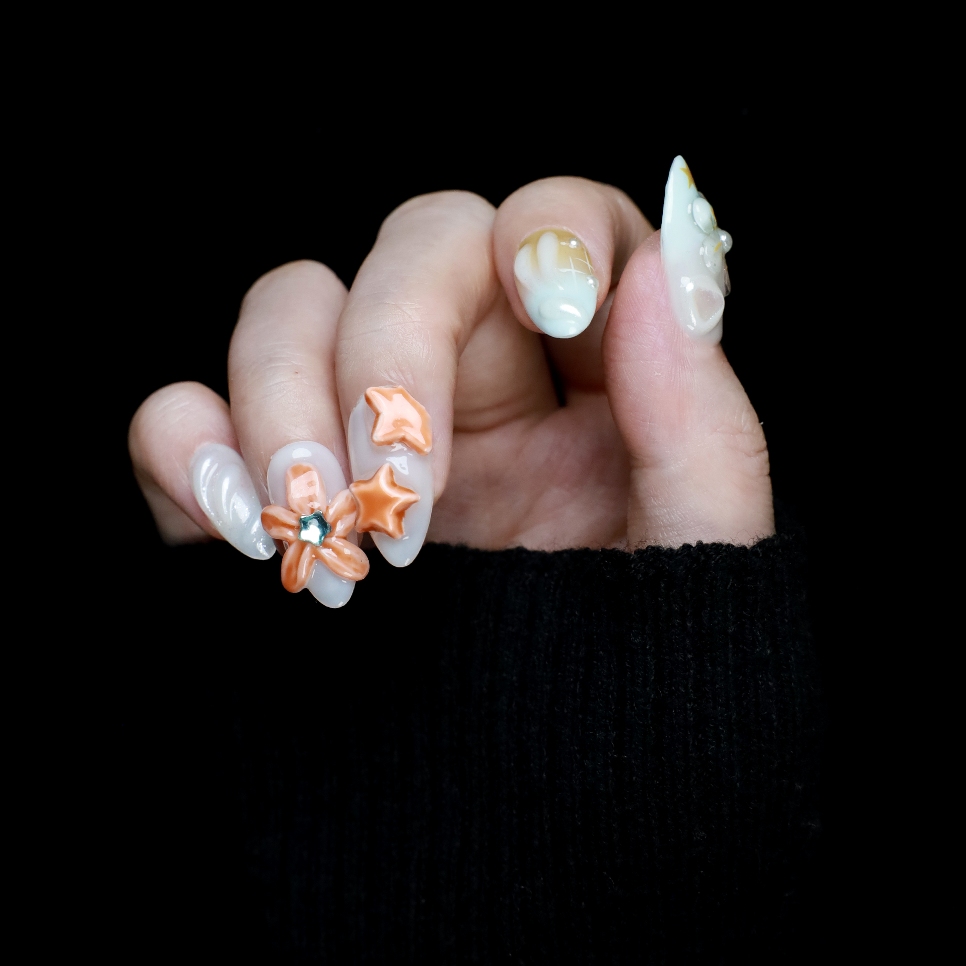 A hand with long, intricately decorated nails exhibits ethereal designs of blue and white swirls, 3D floral accents, pearls, and a metallic silver textured nail against a black background. This look perfectly captures the elegance of We Do Nails Celestial Blossom Press On Nails.