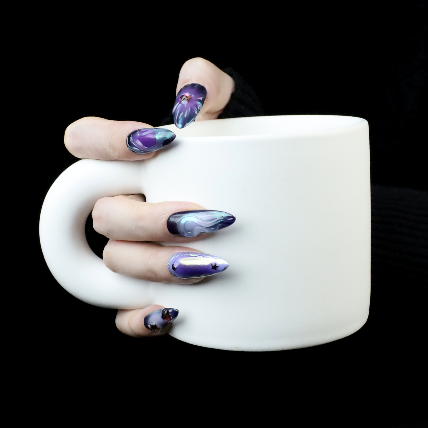A hand with long, artistic purple nails from We Do Nails Egyptian-Inspired Press On Nails set holds a large white mug against a black background. Each nail boasts unique designs in purple and black, exuding luxury and mystique reminiscent of Golden Pharaoh nails.