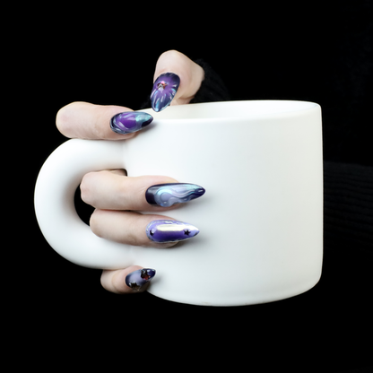 A hand with long, artistic purple nails from We Do Nails Egyptian-Inspired Press On Nails set holds a large white mug against a black background. Each nail boasts unique designs in purple and black, exuding luxury and mystique reminiscent of Golden Pharaoh nails.