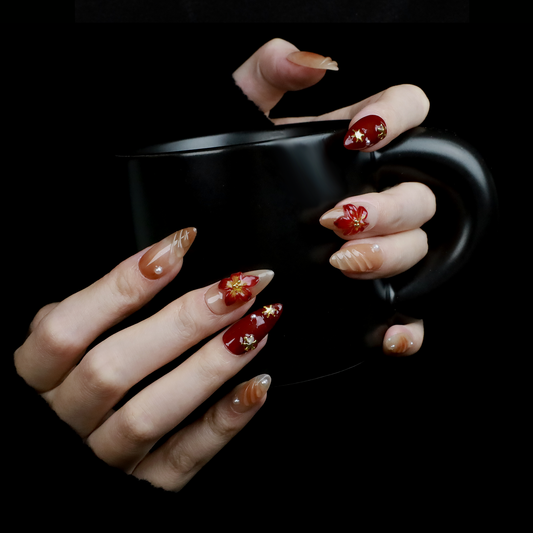 A pair of hands adorned with Elegant Floral Red and Beige Press-On Nails by We Do Nails, featuring intricate red flowers and pearl-gold accents, holds a large black mug. The elongated, almond-shaped nails boast a glossy finish with 3D floral designs against the black background.