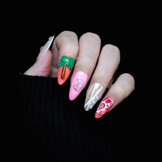 A hand displays the Summer Island Press-On Nails set by We Do Nails, featuring designs like a carrot with leaves, pastel pinks with a jewel, shiny white and silver patterns, and a red and white fish motif. The hand is dressed in a black sleeve against a coordinated backdrop.