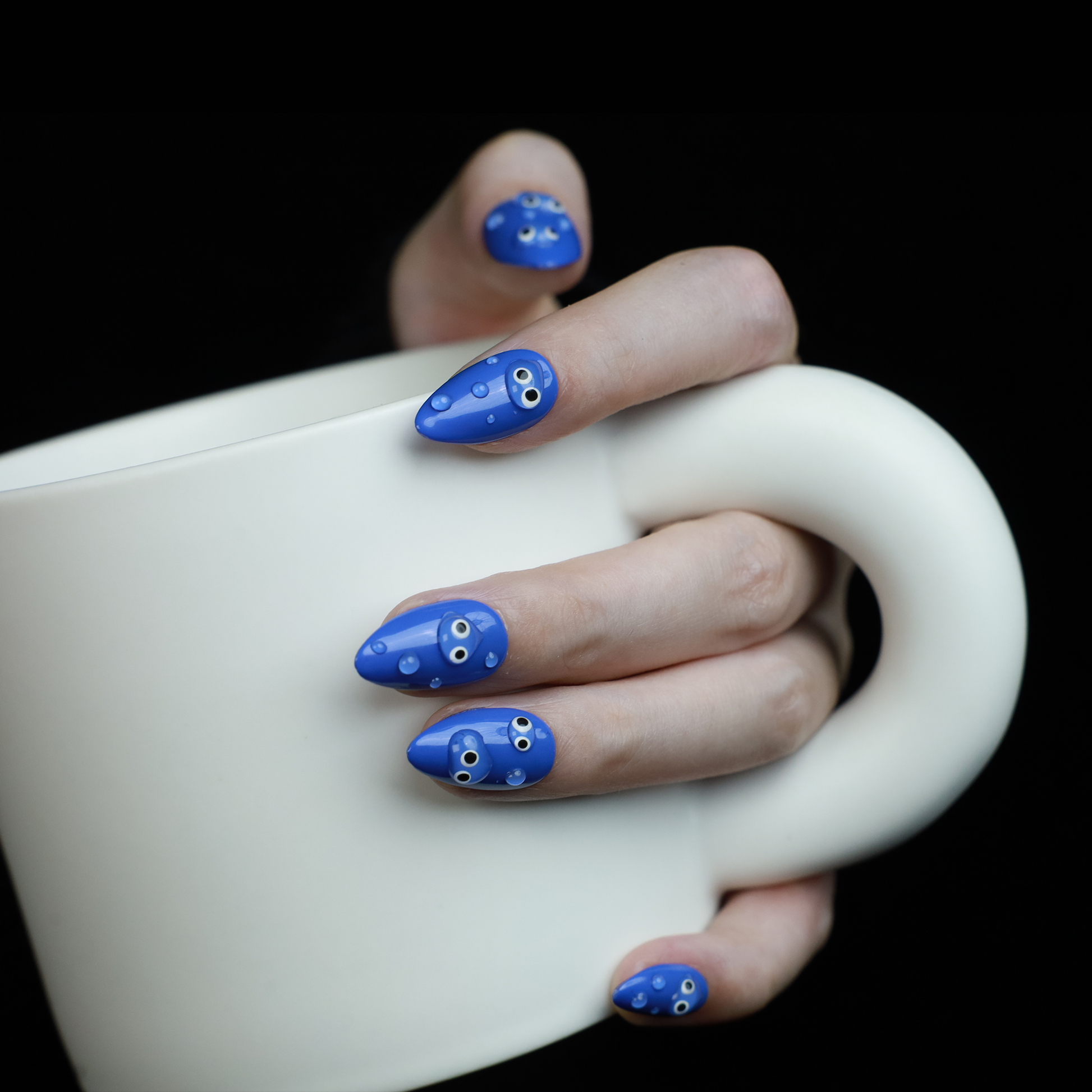 A hand with We Do Nails Cute Blue Monster Press-On Nails set features long, almond-shaped, bright blue nails adorned with playful cartoon faces and 3D googly eyes. The hand in a black sweater is striking against the dark background.