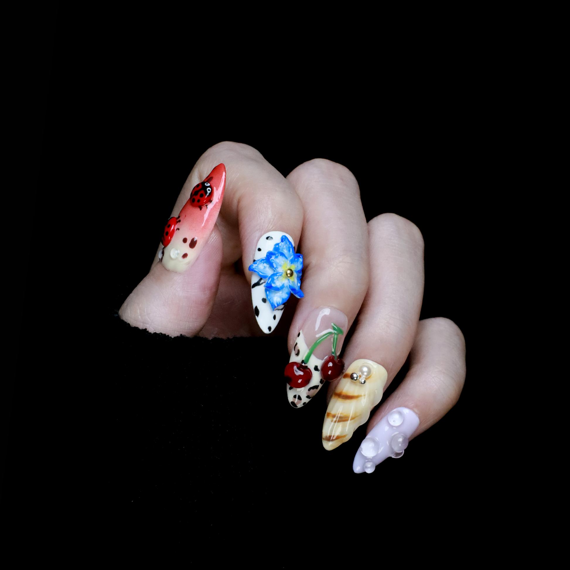The Garden Blooms Press-On Nails by We Do Nails feature a whimsical garden theme with long, decorative nails, including a blue flower, cherries, red and white patterns, pearls, and stripes. Set against a black background, the hand is partially covered by a black sleeve.