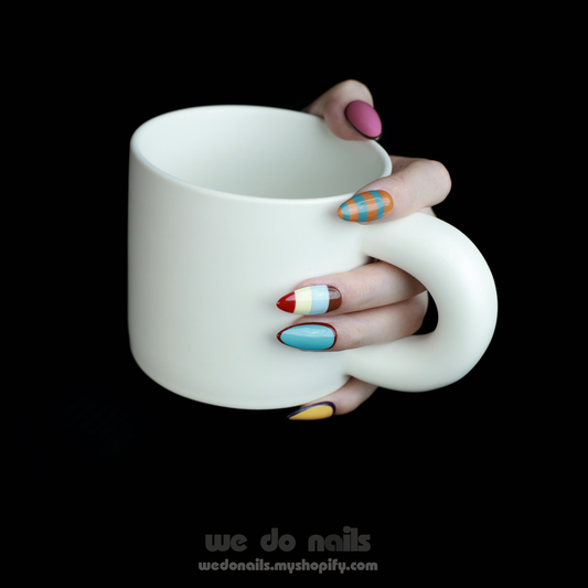 A hand adorned with We Do Nails Vintage Striped Elegance Press-On Nails, featuring a mix of checkerboard, solid hues, and striped patterns in vibrant shades of pink, blue, yellow, red, and white, holds a white mug against a black background.