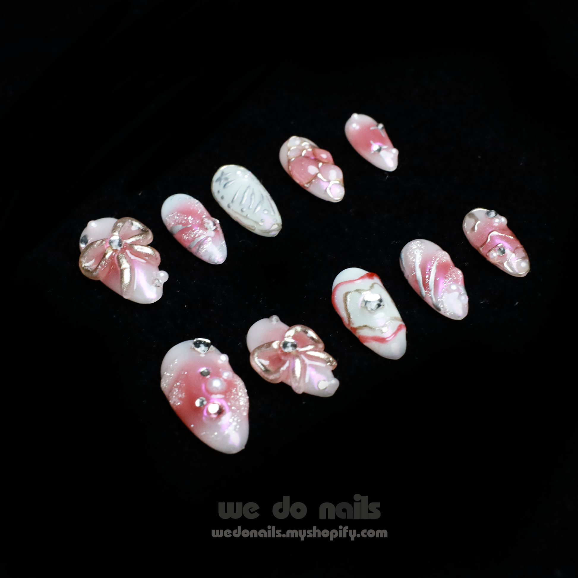A person with intricately designed Blushing Charm Press-On Nails by We Do Nails holds a small, white ceramic handbag. The nails display soft pinks and abstract patterns in pink, white, and gold, accented with shimmering details and decorative embellishments.