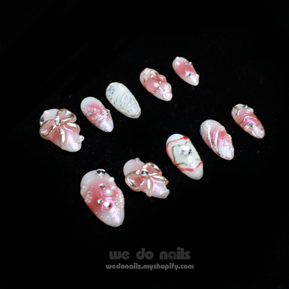 A person with intricately designed Blushing Charm Press-On Nails by We Do Nails holds a small, white ceramic handbag. The nails display soft pinks and abstract patterns in pink, white, and gold, accented with shimmering details and decorative embellishments.
