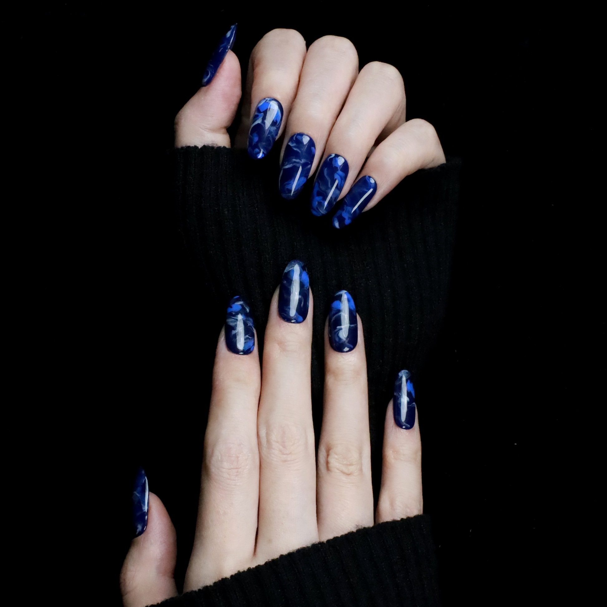 Hands display the Elegant Blue Marble Press-On Nails from We Do Nails, featuring glossy long oval shapes with a blue marble effect. A black ribbed sweater against a black background highlights these striking nails.
