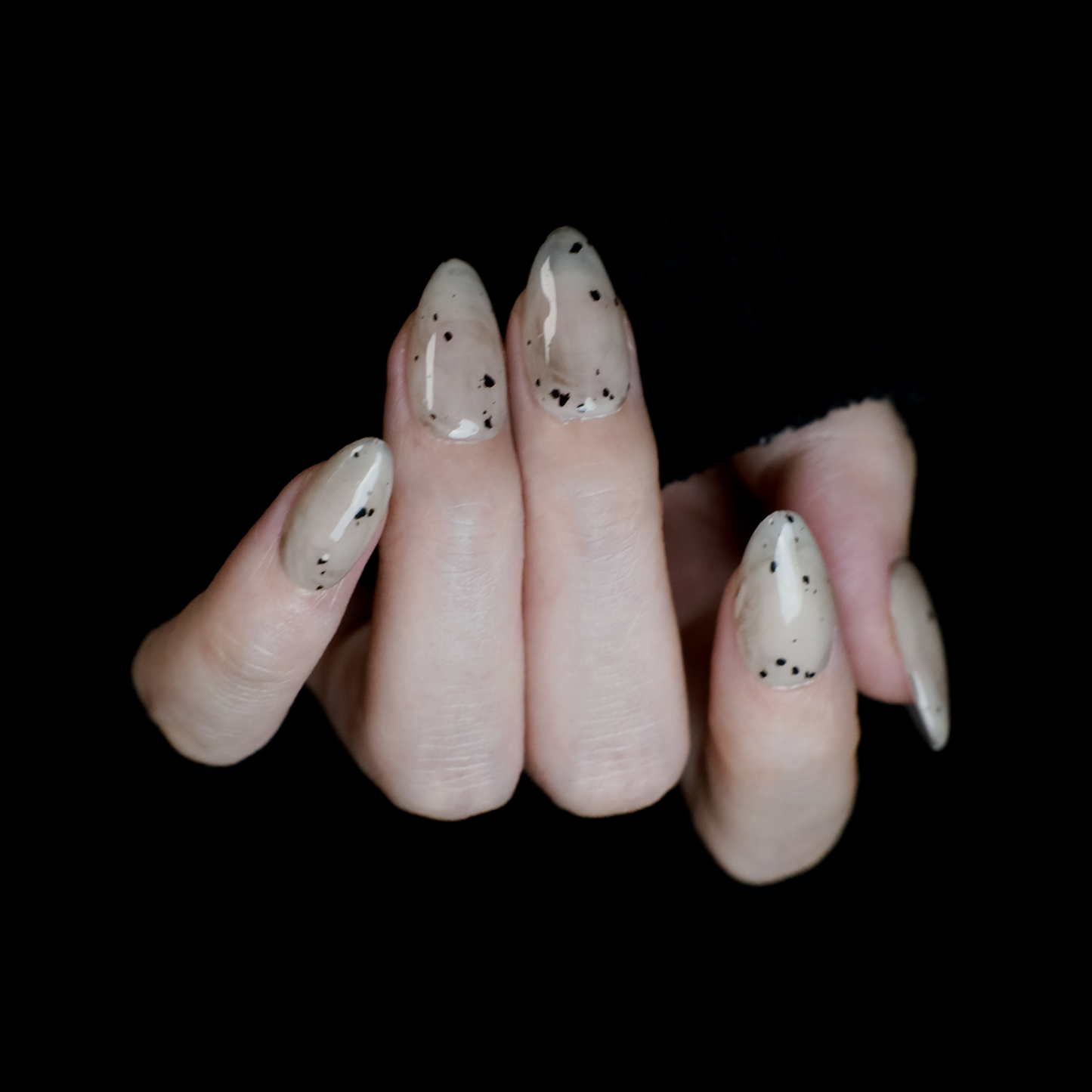 Hands showcasing We Do Nails Marble and Speckled Press On Nails, almond-shaped with a high-gloss shine and scattered speckles, perfectly highlighted against a solid black background.