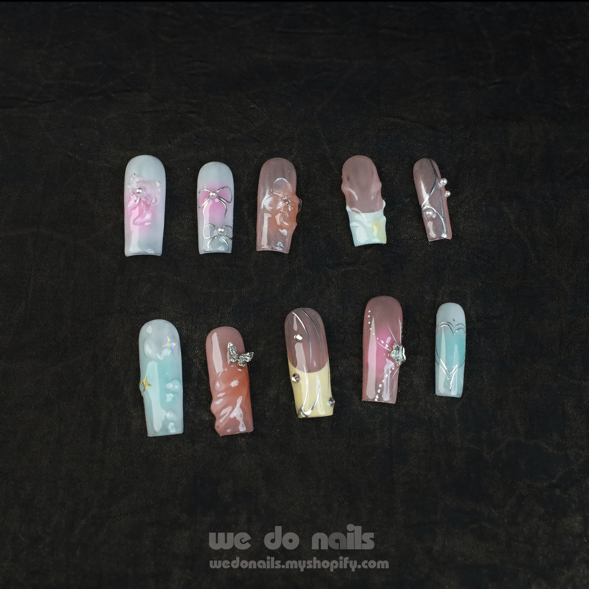 A hand with long, intricately designed nails featuring floral and butterfly designs is posed next to a plain white cylindrical object against a black background. The text We Do Nails wedonails.myshopify.com showcases the vibrant look of our Butterfly Bliss Press-On Nails.