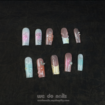 A hand with long, intricately designed nails featuring floral and butterfly designs is posed next to a plain white cylindrical object against a black background. The text We Do Nails wedonails.myshopify.com showcases the vibrant look of our Butterfly Bliss Press-On Nails.