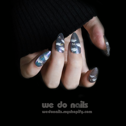 A hand adorned with glittery, pointed Starry Night Press-On Nails featuring eye-catching designs of clouds and stars peeks from a black sleeve. We Do Nails and a website link are displayed at the bottom, against a black backdrop.