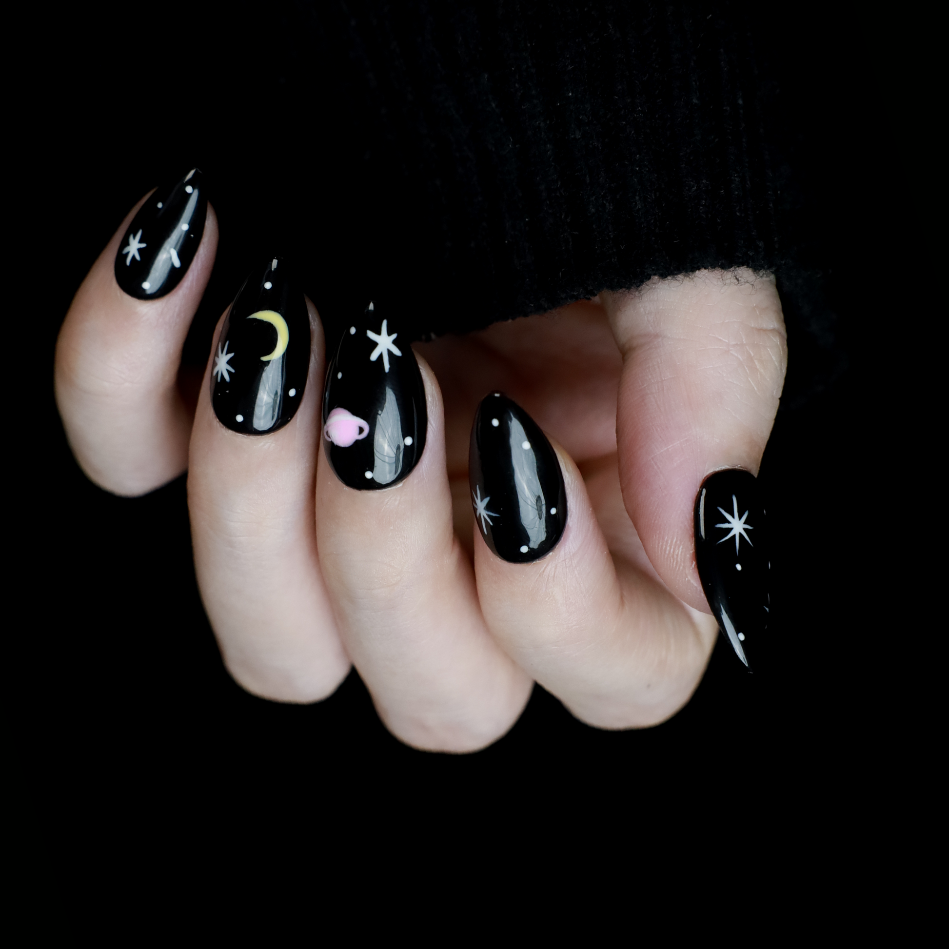 Hands decorated with Galactic Night Sky Press On Nails by We Do Nails, featuring stars, moons, and planets, hold a plain white mug against a dark backdrop.