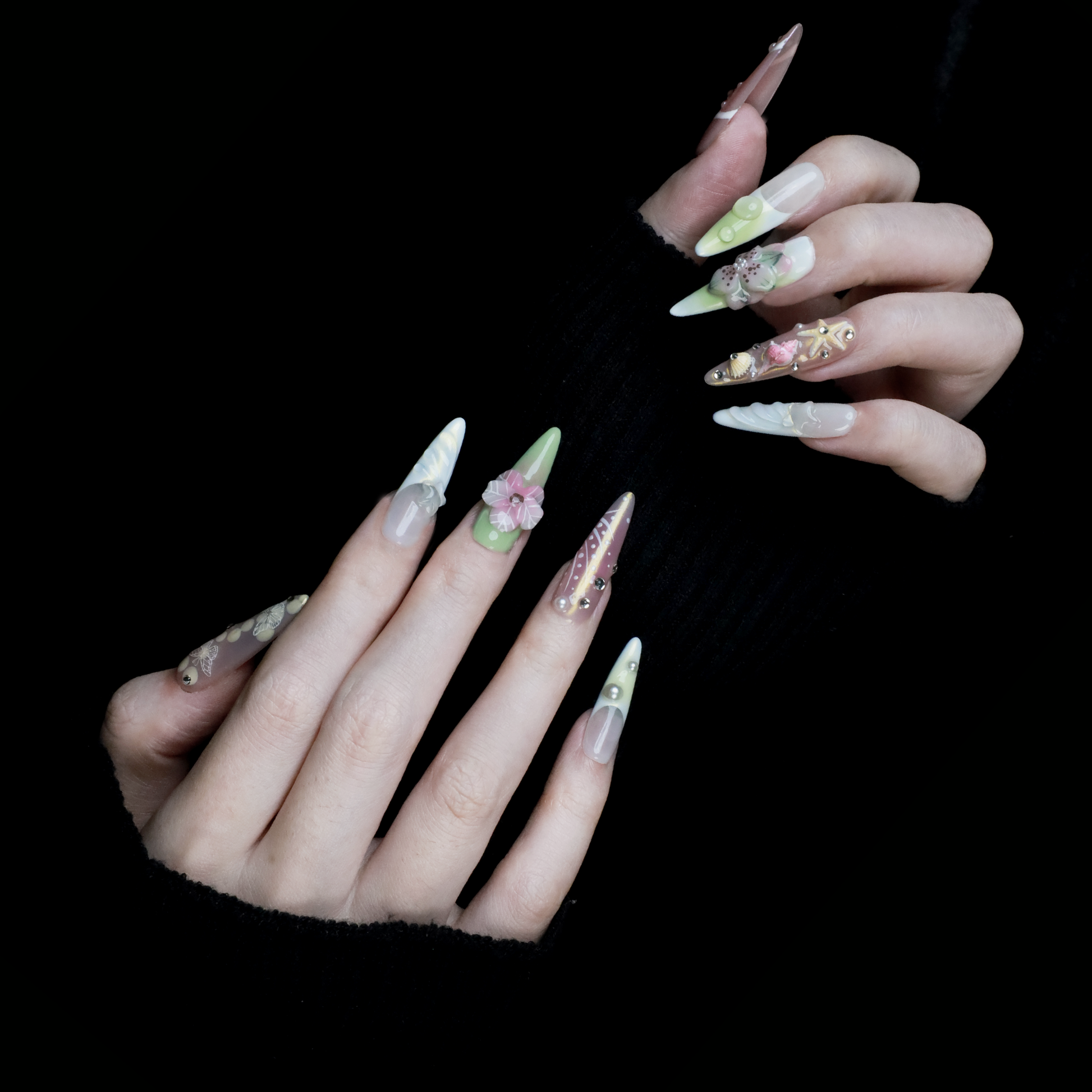 A hand flaunts We Do Nails Floral Blush Pearl Press-On Nails: green with pearls, pink with a butterfly, clear with gold accents, blue with a wave pattern, and a pink-white thumb—all set against a black background for an enchanting look.