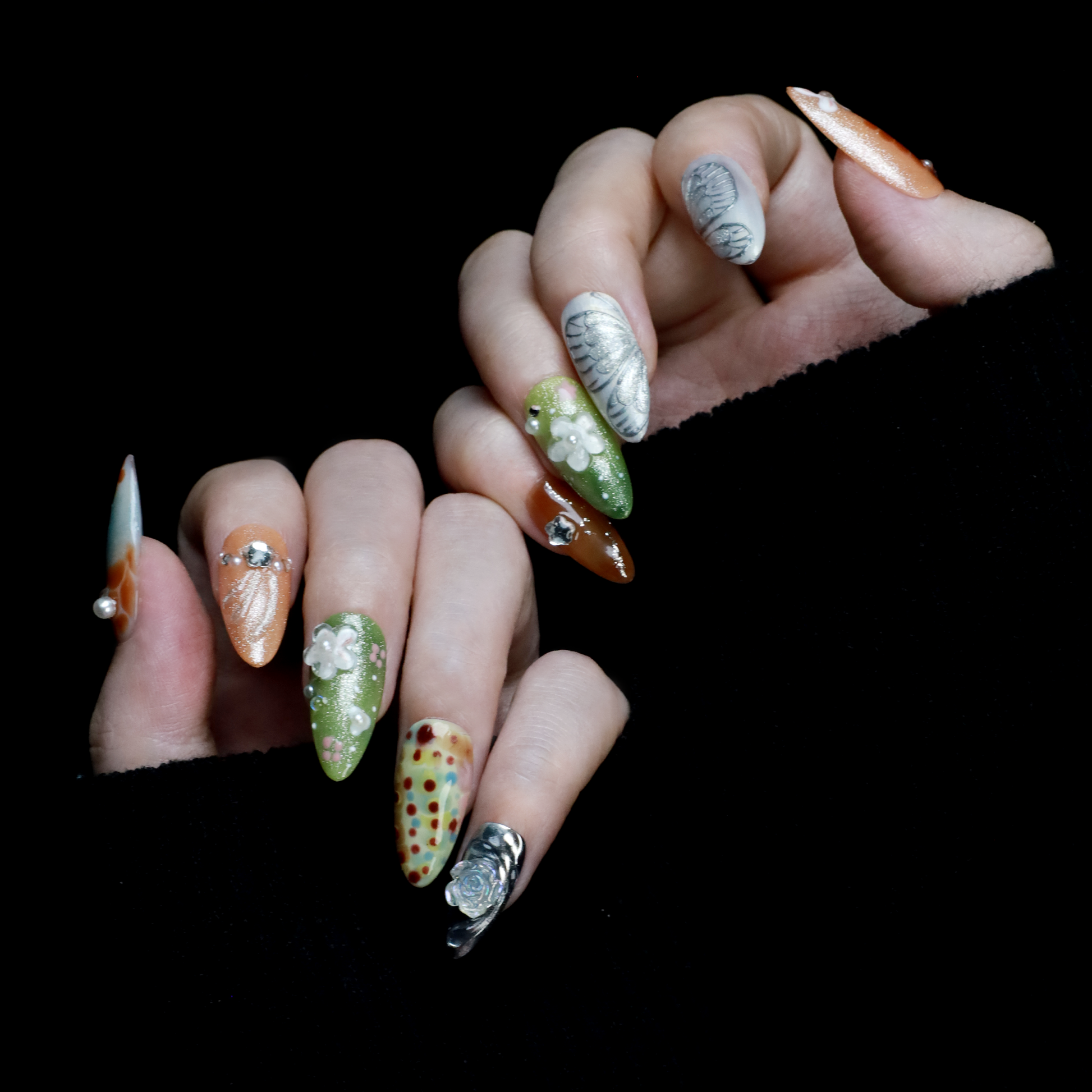 A hand showcases the Fantasy Garden Press-On Nails by We Do Nails against a black background. The nails feature designs of a koi fish with a pearl, peach with rhinestones, 3D green flowers, green with red floral dots, and metallic silver with a crystal accent.