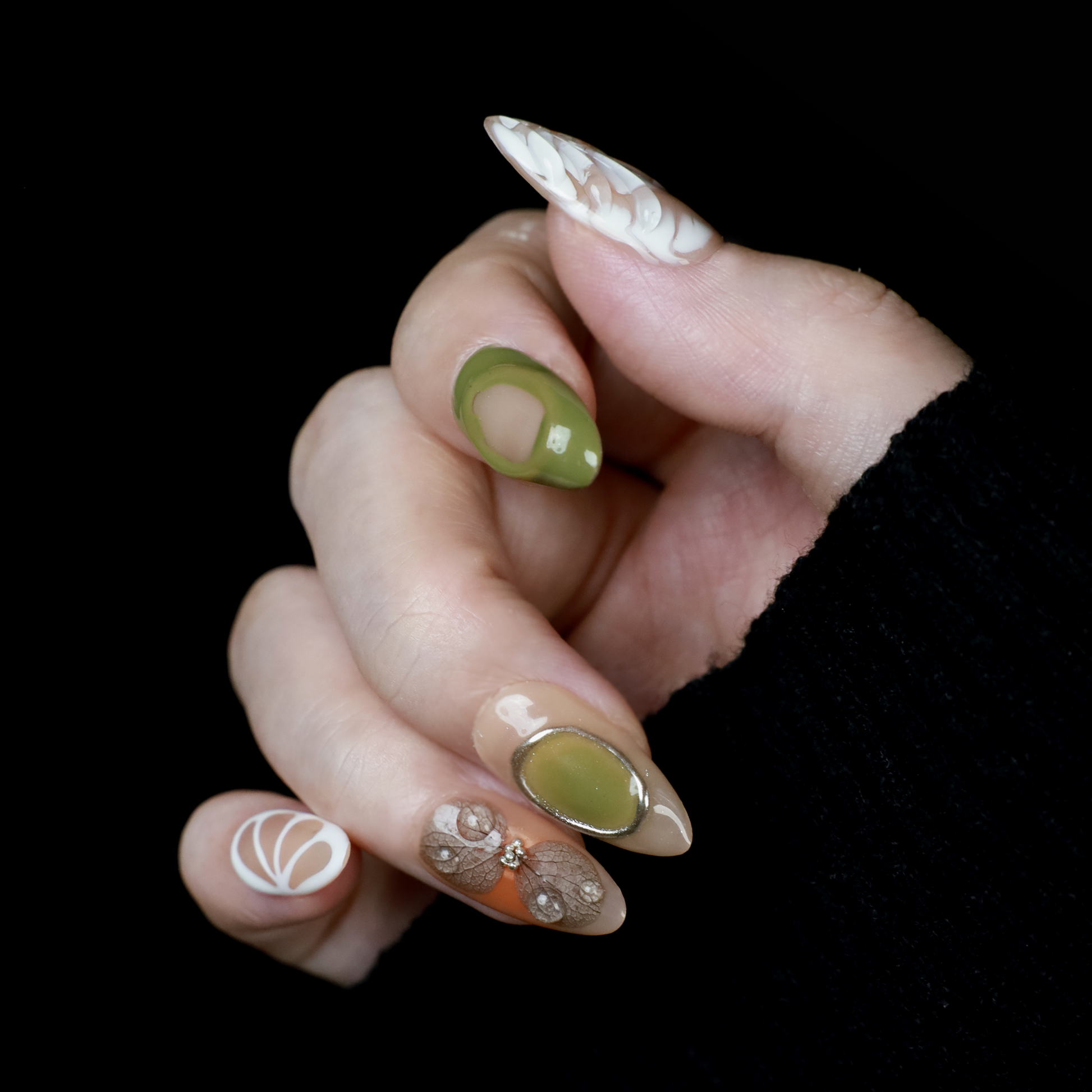A hand in a black sleeve displays the Leaf and Organic design Press-On Nails by We Do Nails, featuring long acrylic nails with white abstract patterns, green accents, and clear 3D embellishments that create a nature-inspired look against the dark background.