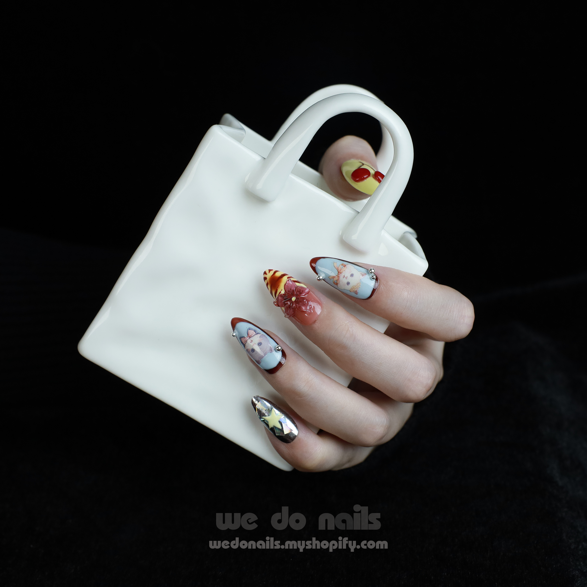 A hand adorned with We Do Nails Extravaganza Press-On Nails, showcasing unique patterns and colors, holds a small white handbag. The dazzling metallics gleam against the black background, highlighting the bold nail art and bag.