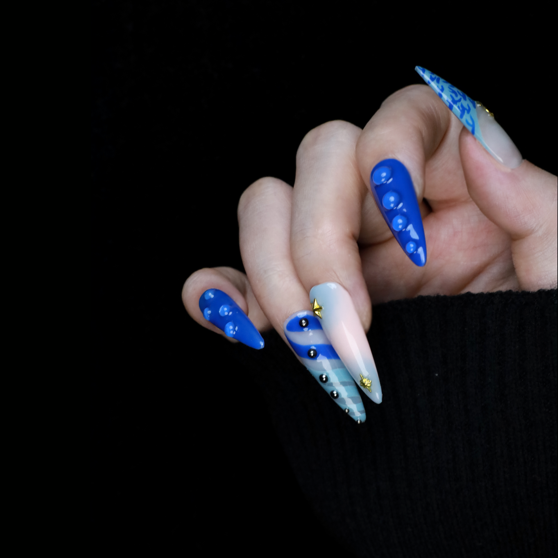 A hand featuring We Do Nails Blue Dreams Press-On Nails with elongated designs and pink hues, includes 3D metallic nail art and textured patterns like ridges and dots against a black backdrop.