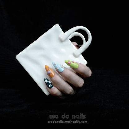 A hand adorned with We Do Nails Pearl Pop Press-On Nails, showcasing colorful patterns and 3D pearl accents, grips a uniquely shaped white mug with two handles. The dark background highlights the intricate nail art and distinctive mug design.