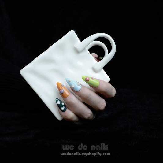 A hand adorned with We Do Nails Pearl Pop Press-On Nails, showcasing colorful patterns and 3D pearl accents, grips a uniquely shaped white mug with two handles. The dark background highlights the intricate nail art and distinctive mug design.