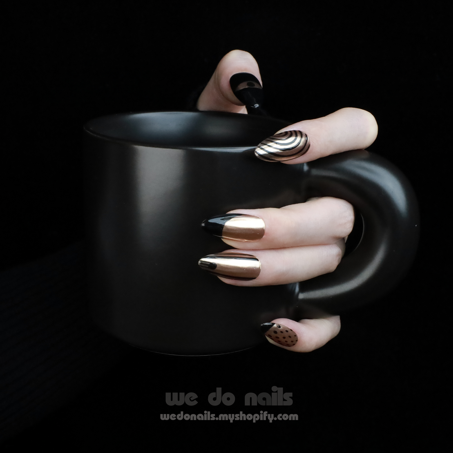 A hand adorned with We Do Nails Egyptian-Inspired Press On Nails in metallic, black, and Golden Pharaoh styles holds a matte black mug. The nails showcase geometric and polka dot patterns inspired by ancient Egypt. A watermark is visible at the bottom of the image.