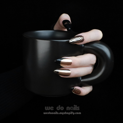 A hand adorned with We Do Nails Egyptian-Inspired Press On Nails in metallic, black, and Golden Pharaoh styles holds a matte black mug. The nails showcase geometric and polka dot patterns inspired by ancient Egypt. A watermark is visible at the bottom of the image.