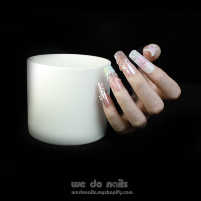 A hand with long, intricately designed nails featuring floral and butterfly designs is posed next to a plain white cylindrical object against a black background. The text We Do Nails wedonails.myshopify.com showcases the vibrant look of our Butterfly Bliss Press-On Nails.