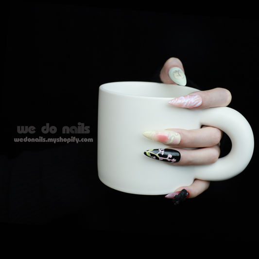 A hand holds a white mug, displaying We Do Nails Abstract Floral Design Press-On Nails, featuring intricate floral patterns in vibrant colors against a dark backdrop. Text: We Do Nails wedonails.myshopify.com.