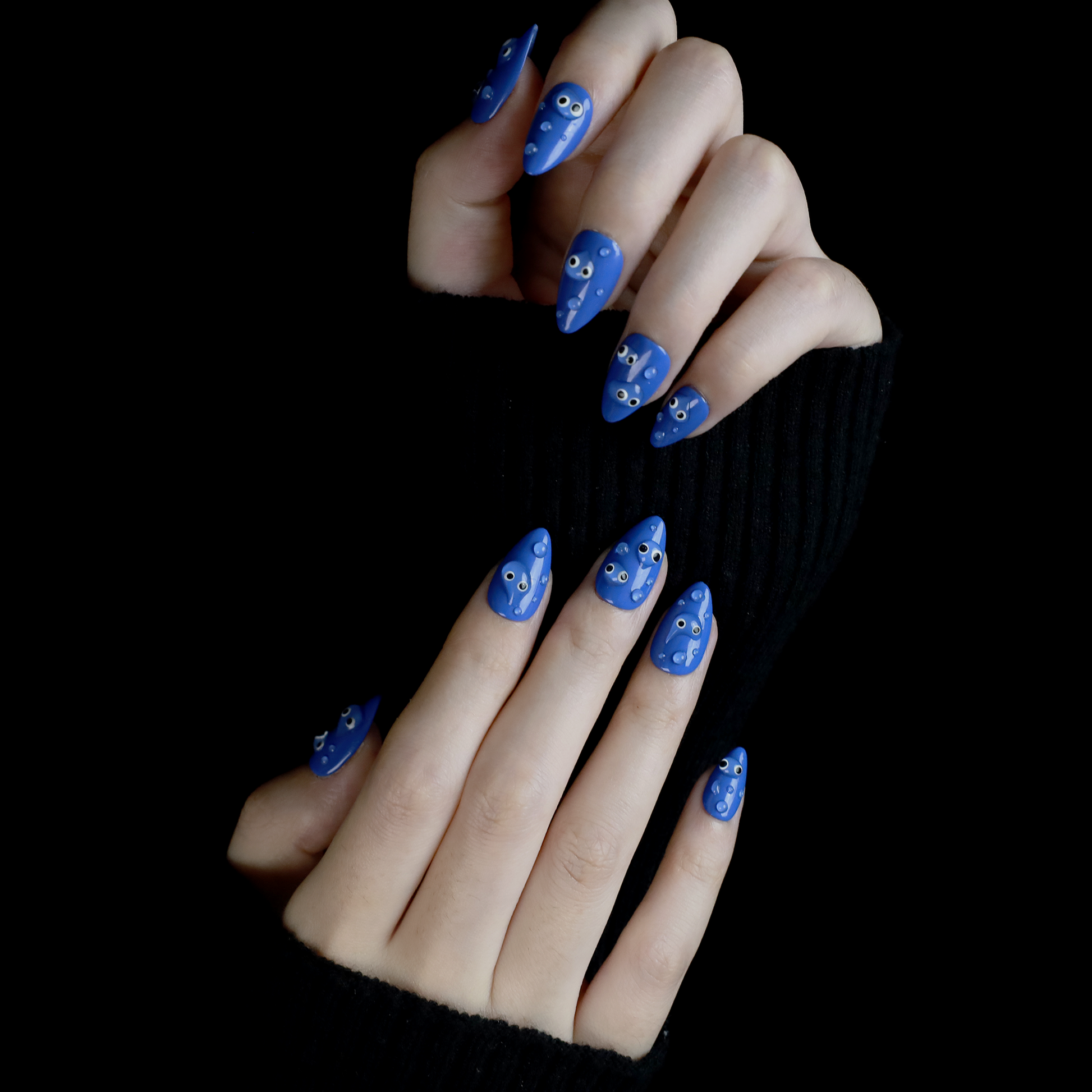 A hand with We Do Nails Cute Blue Monster Press-On Nails set features long, almond-shaped, bright blue nails adorned with playful cartoon faces and 3D googly eyes. The hand in a black sweater is striking against the dark background.