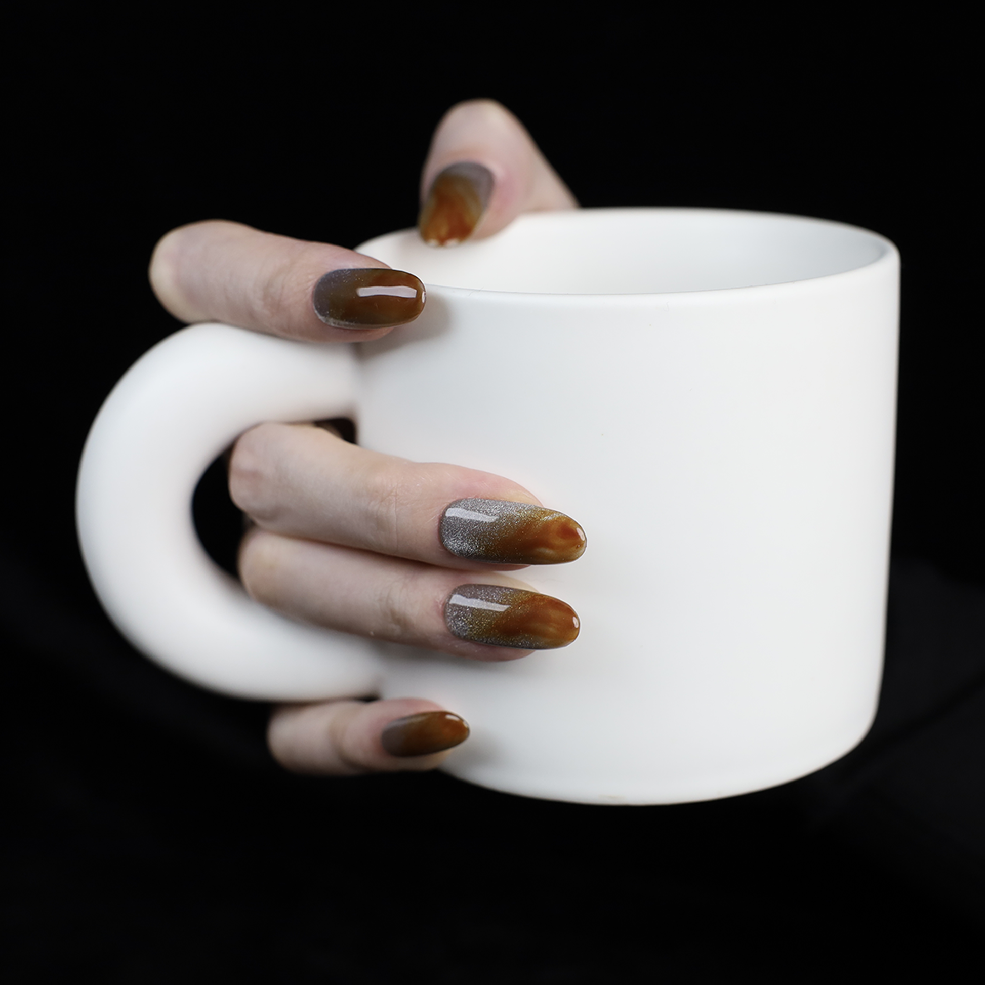 A hand with We Do Nails Elegant Brown Marble Press-On Nails, featuring a subtle metallic chrome accent, holds a simple white mug against a black backdrop.